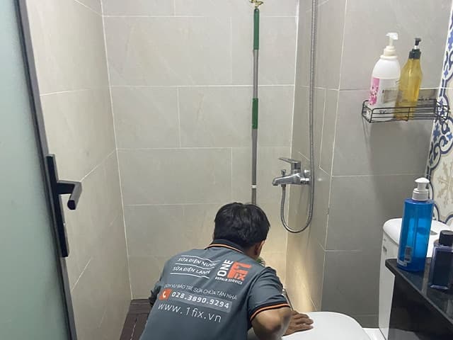 Một bàn tay đang dùng vật nhọn để thông từng lỗ nhỏ trên bề mặt của vòi hoa sen đã được tháo rời.