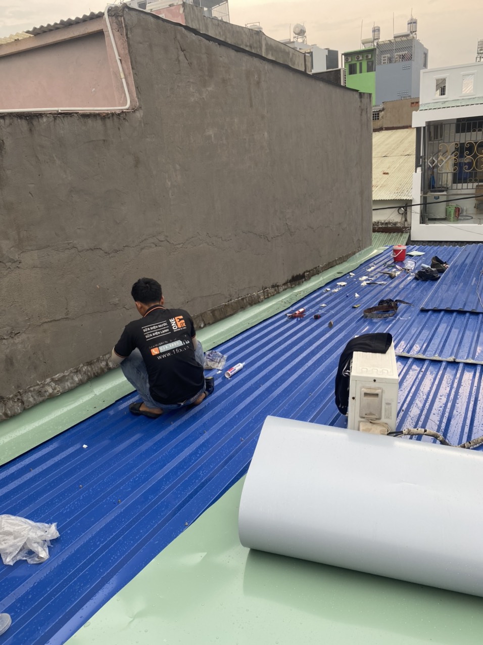 Thợ 1Fix đang thi công sơn chống nóng trên mái tôn — áo đồng phục 1Fix, mái tôn đã phủ lớp sơn xanh chống nóng, cuộn xốp cách nhiệt bên cạnh
